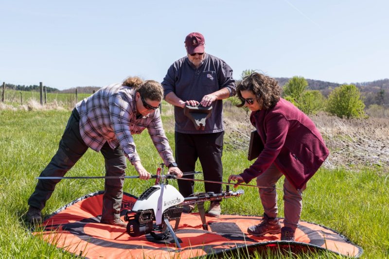 A student and faculty member are touching a drone on the ground placed on an orange, circular helicopter pad while another faculty member stands behind holding the control pad and presses a button.