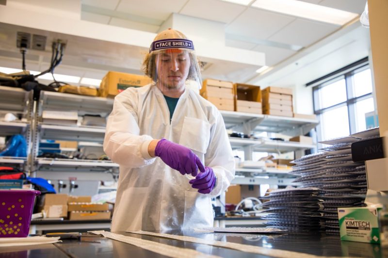  A student wearing a plastic face shield, lab coat, and purple medical gloves looks down at lab materials as they hold onto a transparent, plastic container with a liquid inside.