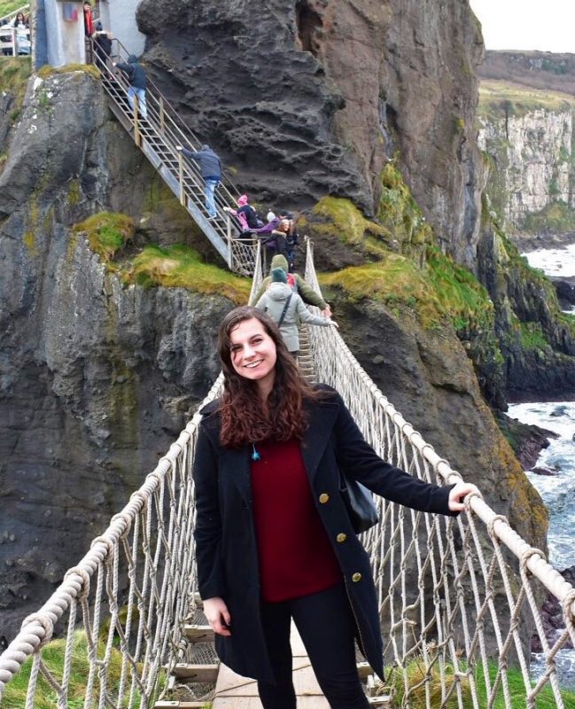 Student stands on a swinging bridge with a mountain in the background and the ocean underneath