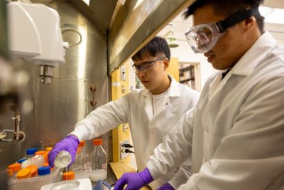 two students in white lab coats and eye protection in a lab